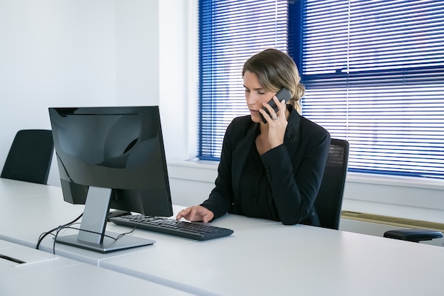 사무실에서 직장에서 컴퓨터를 사용하는 동안 휴대 전화에 얘기하는 소송에서 여성 비즈니스 리더를 집중. 미디엄 샷. 디지털 커뮤니케이션 및 멀티 태스킹 개념