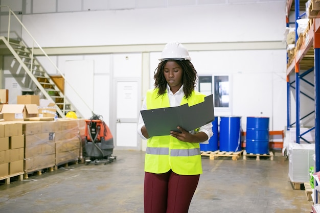 Free photo focused female logistic employee in hardhat and safety vest walking in warehouse, carrying open folder, looking through document. copy space, front view. labor and inspection concept
