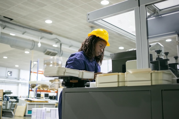 Focused female paper factory employee wearing hardhat, operating industrial machine, standing at control board