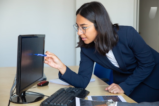 Сфокусированный женский профессиональный указывая ручкой на статистический отчет на мониторе, опираясь на офисный стол с финансовыми диаграммами. Средний план. Концепция финансового консультанта