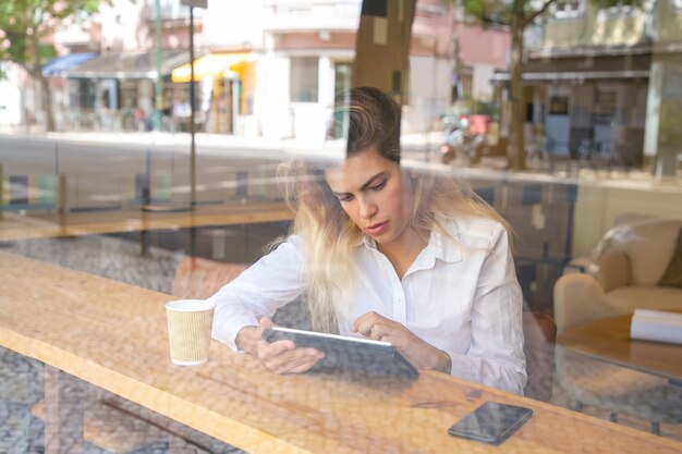 Focused female professional sitting at desk in co-working space or coffee shop, using tablet