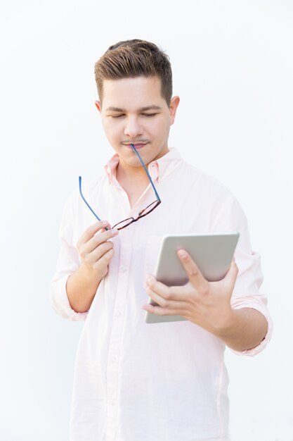 Focused guy biting eyeglasses, reading on tablet screen