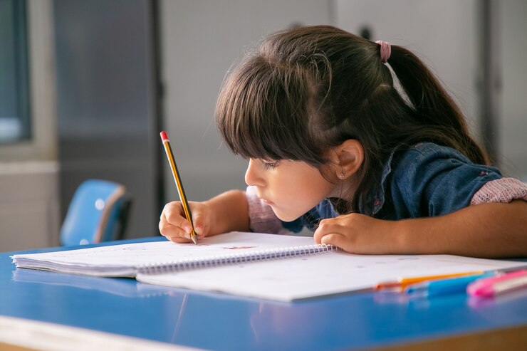 Free Photo focused haired latin girl sitting at school desk and drawing in her copybook