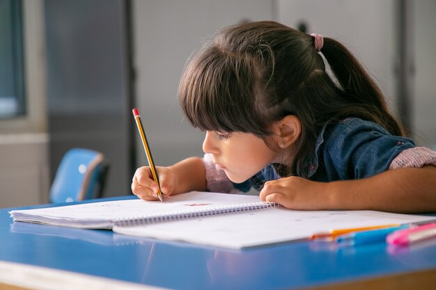 学校の机に座って、彼女のコピーブックに描く焦点を絞った髪のラテンの女の子