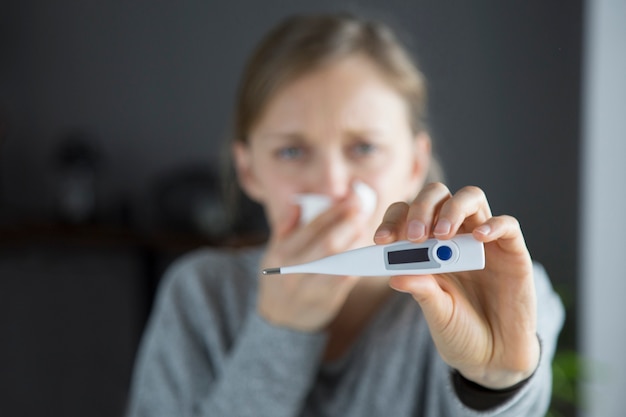 鼻をかむ病気の女性によって保持されている温度計のイメージを集中