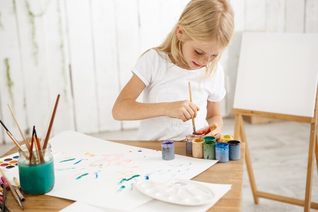 Focused and inspired little blonde girl deeping brush into paint, mixing it. Female freckled child in white t-shirt occupied with painting.