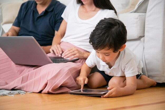 Focused little boy using tablet by himself, sitting on floor in living room by his parents with laptop.