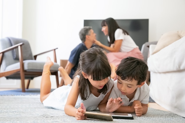 거실 바닥에 누워 부모가 키스하는 동안 학습 앱으로 디지털 기기를 사용하는 집중된 어린 아이들