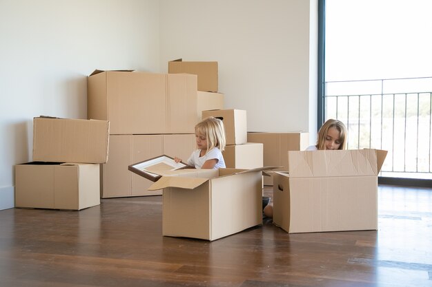 Focused little kids unpacking things in new apartment, sitting on floor and taking objects from open cartoon boxes