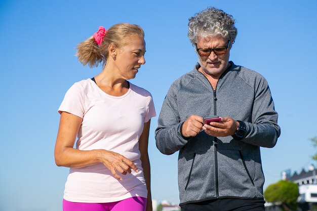 ジョギング後に携帯電話のフィットネスアプリを使用して集中した男性ジョガー。スポーツ服を着て、外に立っている成熟したカップル。スポーツコンセプトのためのコミュニケーションとガジェット