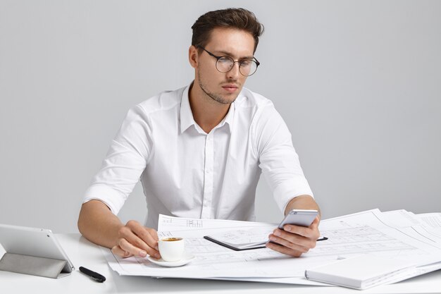 Focused male office worker uses smart phone for online communication, drinks espresso or cappuccino, sits at work place, has serious expression. Young man works on architectual project alone