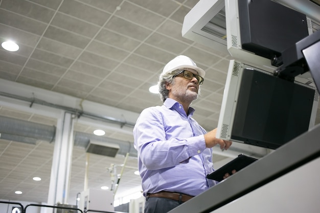 Free photo focused male plant engineer in hardhat and glasses operating industrial machine, pushing buttons on control panel