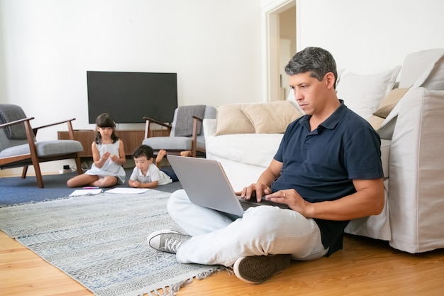 Focused man working at home, sitting on floor and using laptop while two kids drawing