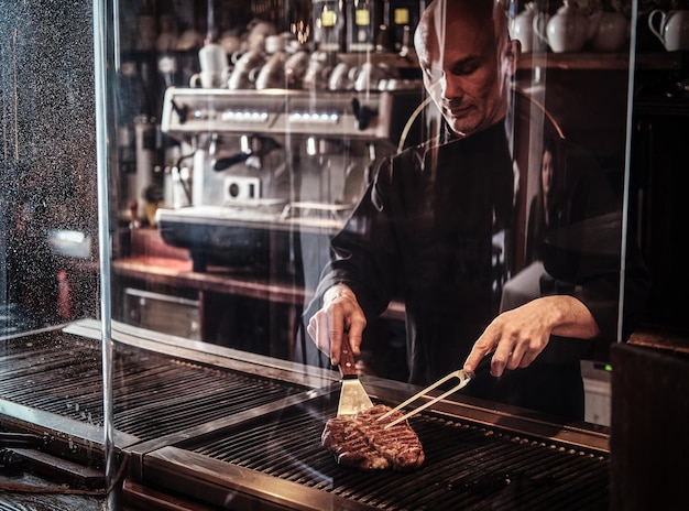 Сосредоточенный шеф-повар готовит вкусный стейк из говядины на кухне, стоя за защитным стеклом в ресторане.