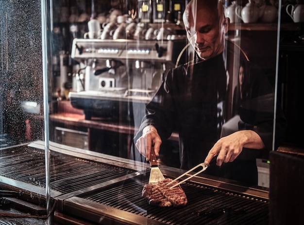 Focused master chef cooking delicious beef steak on a kitchen, standing behind protective glass in a restaurant.