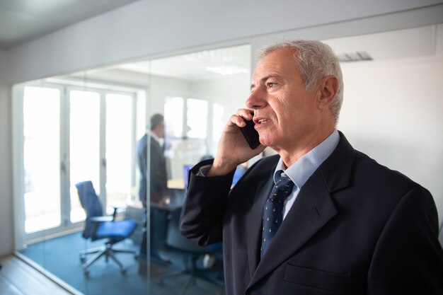 Сфокусированные зрелые бизнесмен разговаривает по мобильному телефону на офисной стеклянной стене, стоя в коридоре. Концепция коммуникации