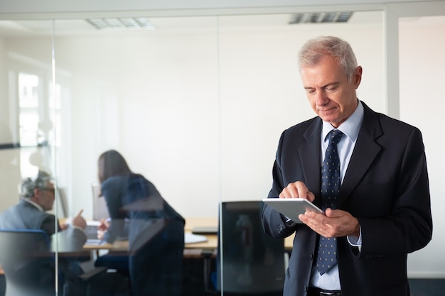 Focused mature businessman using tablet while his colleagues discussing project at workplace behind glass wall. Copy space. Communication concept