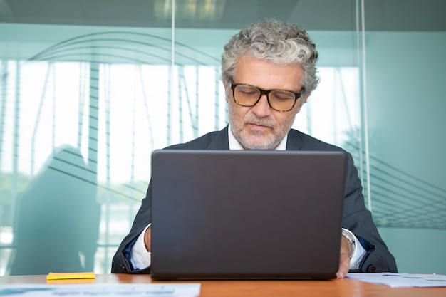 Focused mature professional wearing suit and glasses, working at computer in office, using laptop at table