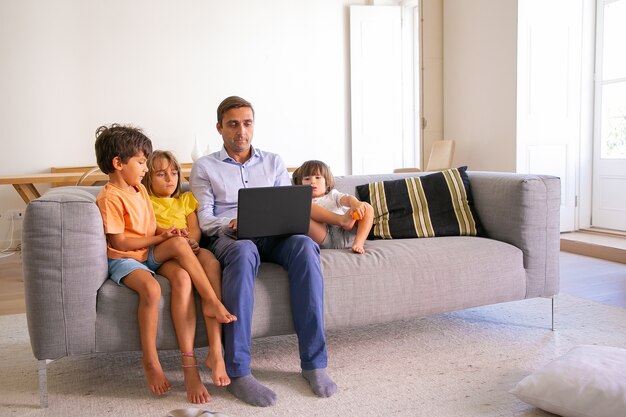 Focused middle-aged dad sitting on sofa with kids and typing on laptop. Caucasian father relaxing with cute children in living room and watching movie. Digital technology and fatherhood concept