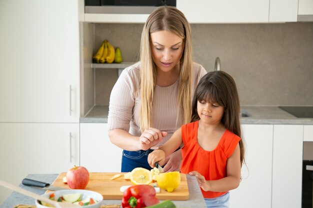 자르고 보드에 신선한 야채를 절단하는 그녀의 소녀를보고 집중된 엄마. 저녁 식사를 요리하는 어머니를 돕는 아이. 가족 요리 함께 개념