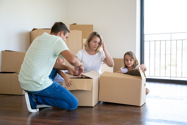 Focused parents and kids unpacking things in new apartment, sitting on floor and opening boxes