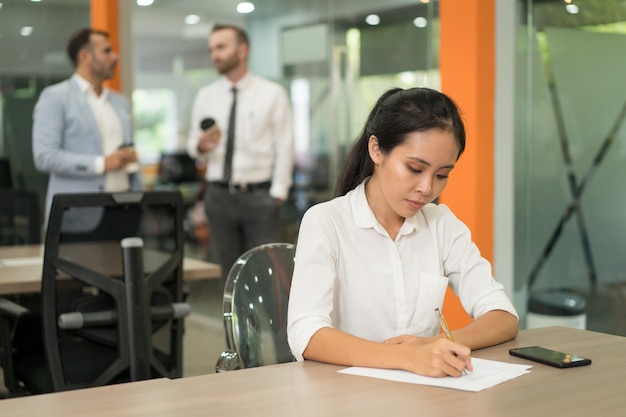 Focused pretty Asian business woman writing at desk in office