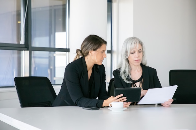 Focused professionals analyzing reports together. Two businesswomen sitting together, reading documents, using tablet and talking. Teamwork concept