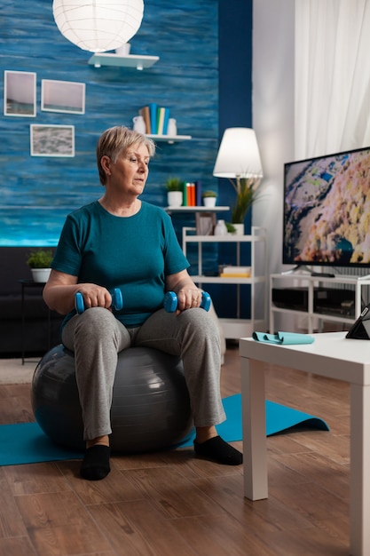 Focused senior woman doing arm exercising using workout dumbbells sitting on swiss ball