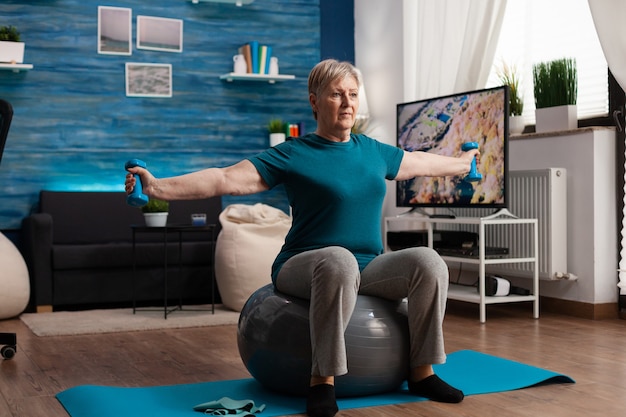 Free photo focused senior woman stretching arm working at body muscle using fitness dumbbells sitting on swiss ball in living room. caucasian male exercising muscular healthcare during wellness workout