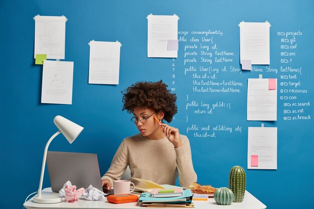 Focused serious Afro American woman looks attentively at screen of laptop computer, works on research project online