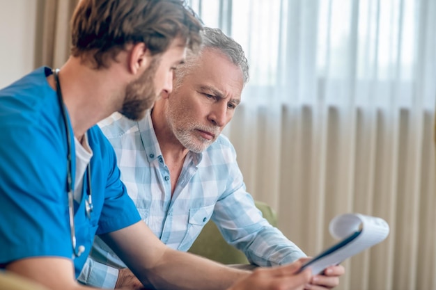 Focused serious gray-haired man in a plaid shirt looking a clipboard in the doctor hands