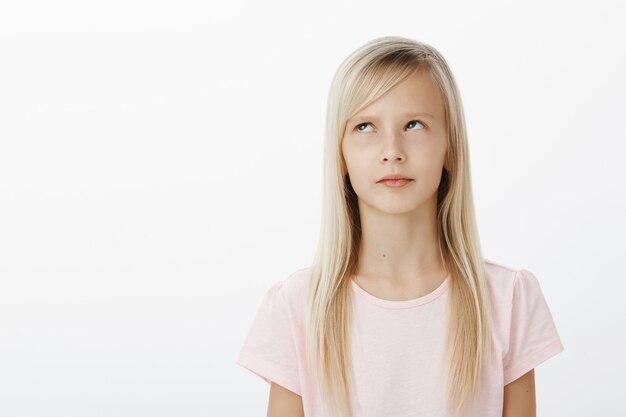 Focused serious young kid recalling learned material while answering near blackboard at school. concerned thinking cute girl, looking up and standing over gray wall remembering