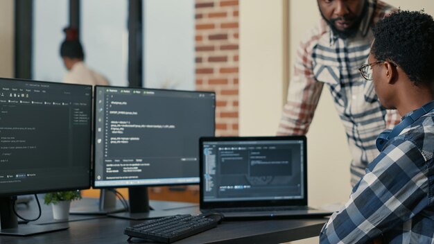 Focused software developer writing code on laptop looking at multiple screens with programming language is interrupted by colleague coder asking for advice. Programmers doing online cloud computing.