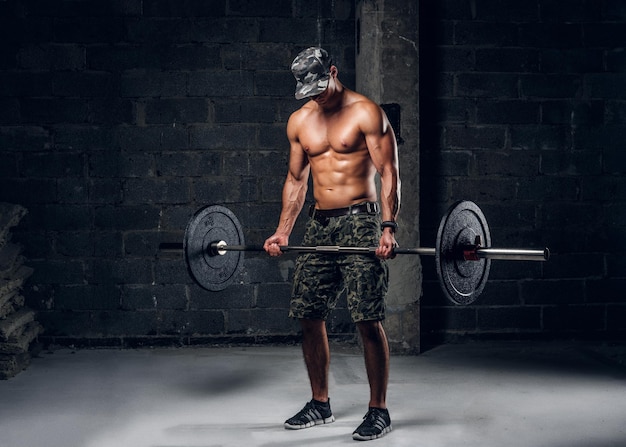 Focused strong man in cap is lifting barbell at dark photo studio.