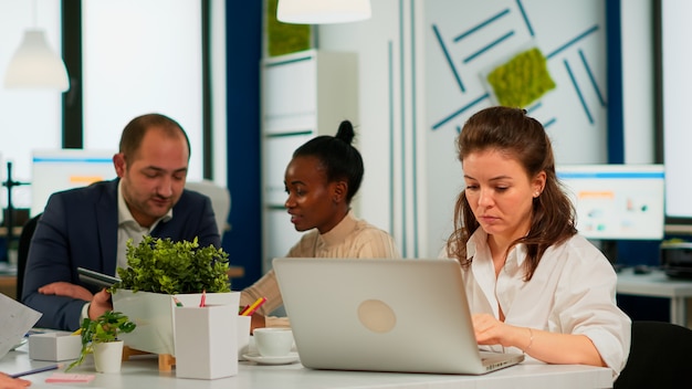 Focused woman manager typing on laptop, browsing on internet while sitting at desk concentrated having multitasks. Multiethnic coworkers talking about startup financial company in modern office.