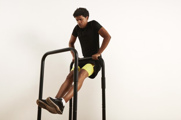 Сосредоточенный молодой афро-американский спортсмен в черной спортивной одежде выполняет тяги собственного веса на мобильных барах, изолированных на белом