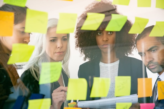 Focused young businesspeople watching at stickers and making notes. Successful concentrated colleagues in suits meeting in office room. Teamwork, business and brainstorm concept