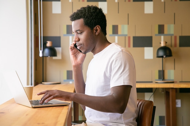 Focalizzato giovane imprenditore che lavora al computer portatile e parlando al telefono cellulare in uno spazio di co-working