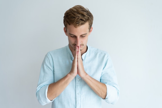 Focused young man praying and keeping hands together. Guy asking God for something. 