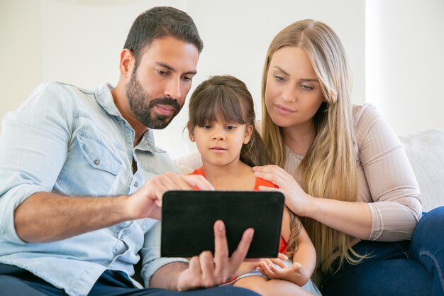 Focused young parents and cute daughter sitting on couch, using tablet, staring at screen, watching video together.