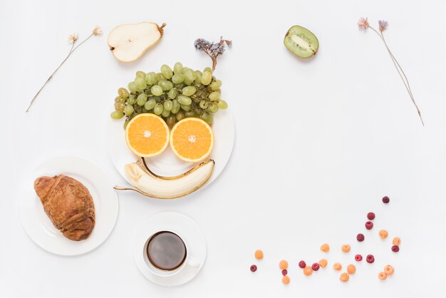 Food arranged as human face on plate with coffee; croissant and coffee on white background