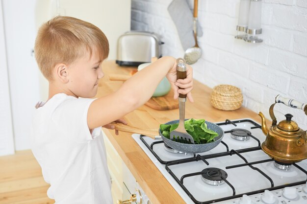 Food, children and cooking concept. Portrait of handsome schoolboy in white t-shirt standing at kitchen counter using stove to cook dinner, holding metal turner, stewing green leaves on frying pan