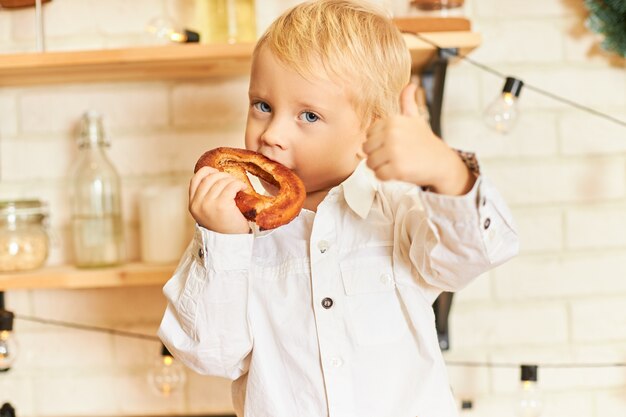 Еда, кулинария, кондитерские изделия и хлебобулочные концепции. Портрет красивого голубоглазого маленького мальчика, наслаждающегося свежеиспеченным бубликом во время завтрака на кухне, жестикулирующего, делая знак пальца вверх
