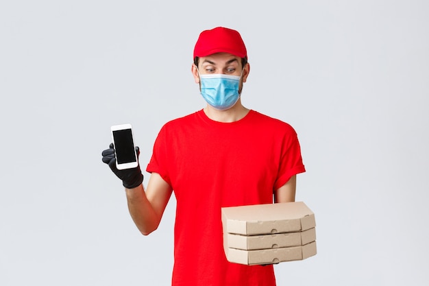 Food delivery, application, online grocery, contactless shopping and covid-19 concept. Excited courier in red uniform, looking at pizza boxes amused, showing smartphone screen app or bonus promo