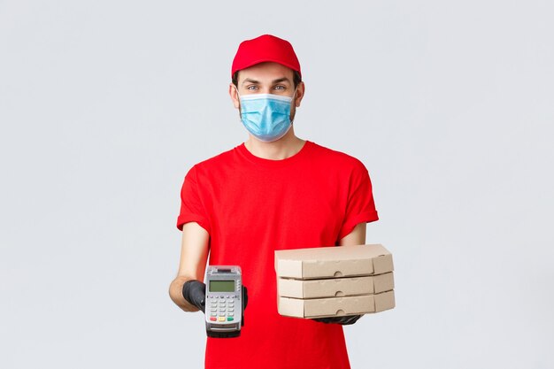 Food delivery, application, online grocery, contactless shopping and covid-19 concept. Friendly courier in red uniform, face mask and gloves, holding order pizza boxes and give client POS terminal