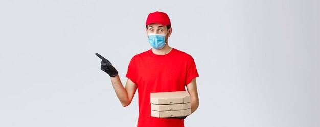 Food delivery application online grocery contactless shopping and covid19 concept Courier brings foor order to client house Delivery guy in red uniform face mask and gloves pointing left