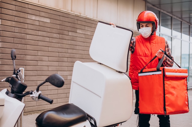 Ragazzo delle consegne di cibo alla guida di uno scooter con scatola con cibo e maschera da indossare