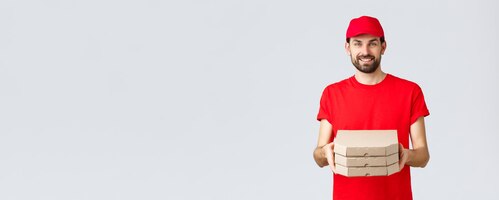 food delivery quarantine stay home and order online concept smiling nice bearded courier in red uniform cap and tshirt handing clients boxes with pizza deliver order grey background