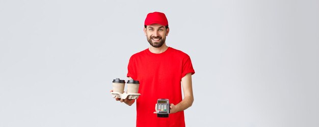 Food delivery quarantine stay home and order online concept Friendly bearded courier in red uniform handing coffee delivery and POS terminal for client to pay contactless grey background