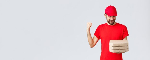 Food delivery, quarantine, stay home and order online concept. Happy, excited bearded courier in red t-shirt and cap celebrating, rejoicing, looking at pizza orders, grey background.